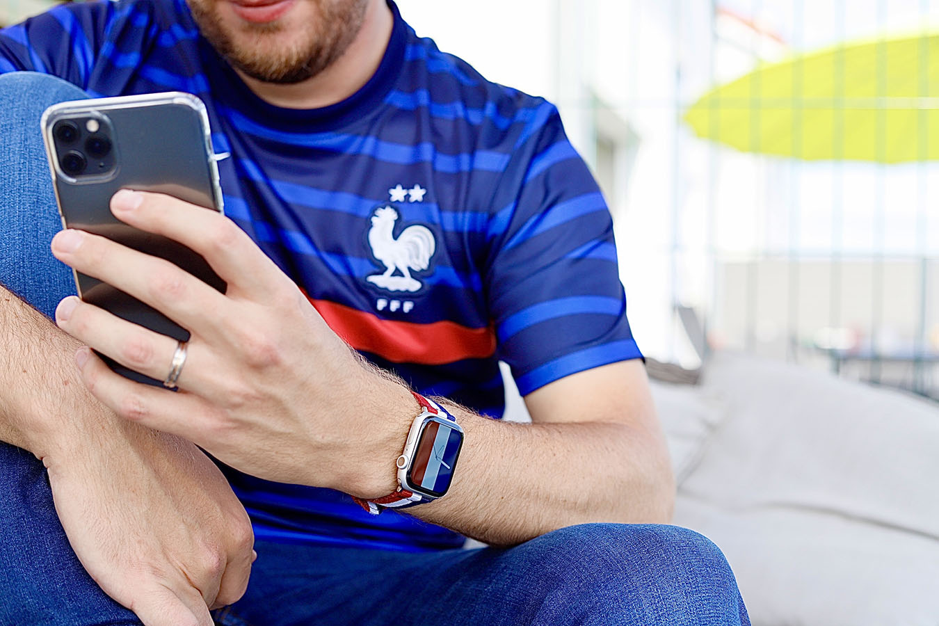 Jeune homme qui porte un maillot de l'équipe de France de football avec un iPhone dans la main et une Apple Watch avec un bracelet aux couleurs du drapeau français. ©2021 Mathieu Improvisato